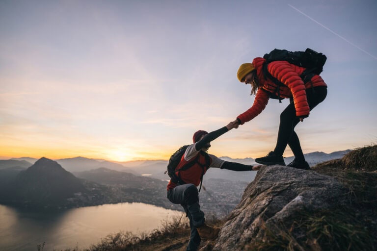 Eine Wanderin hilft einem anderen Wanderer beim Aufstieg auf einen Felsen, im Hintergrund sind Berge, ein See und der Sonnenuntergang zu sehen.