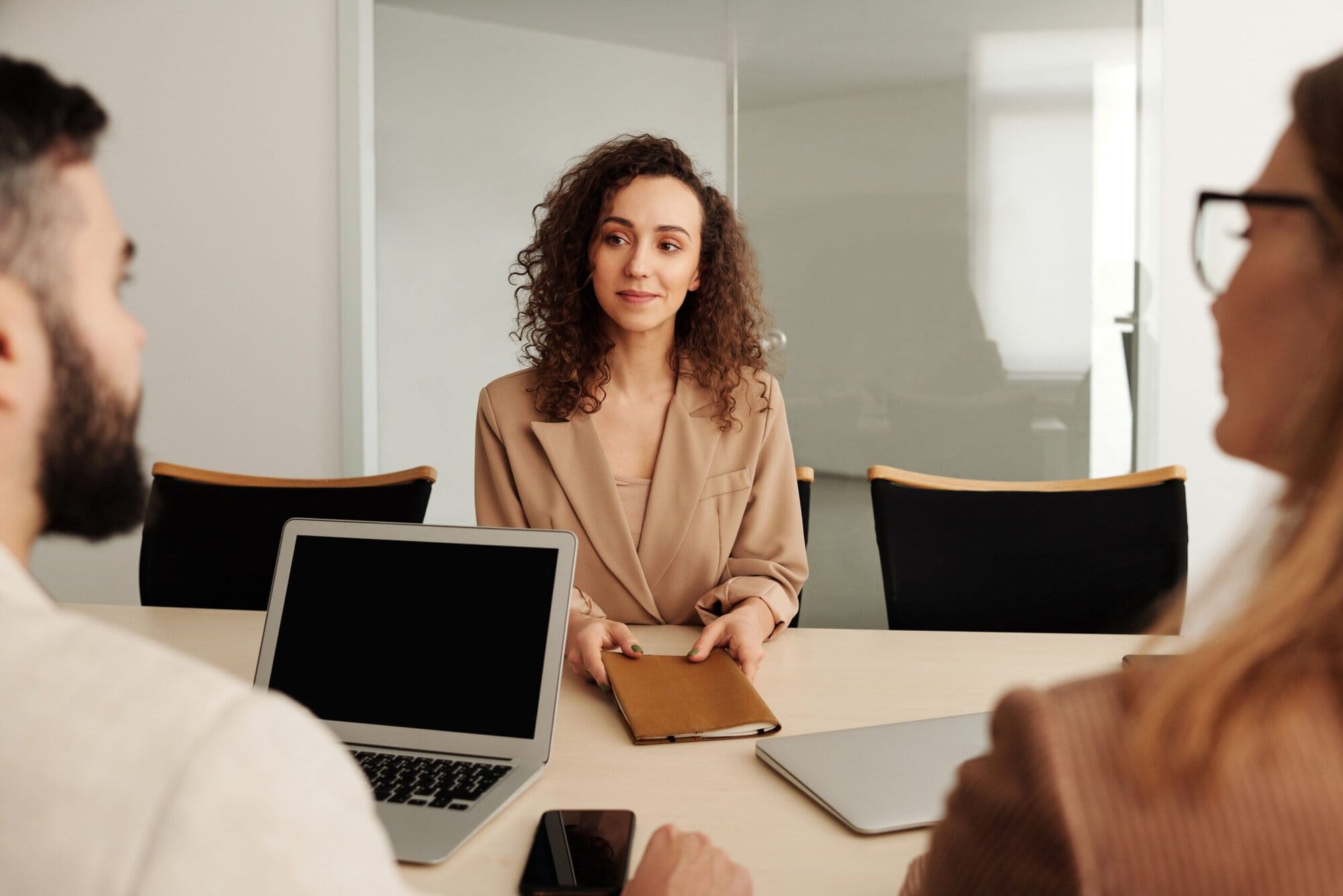 Frau bei einem Gespräch im Büro; ihr gegenüber sitzen zwei Personen mit Laptop und Unterlagen am Konferenztisch.